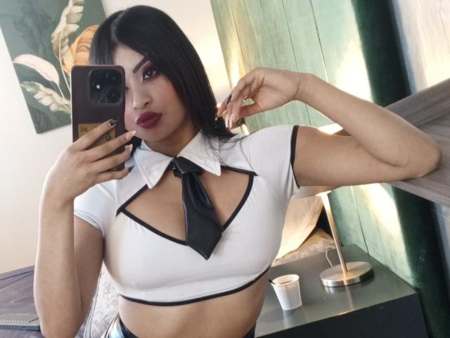 EmmaaRodriguez with dark hair, red lipstick, and a white crop top with black trim and necktie, showing her tits and holding a phone, in a mirror selfie.