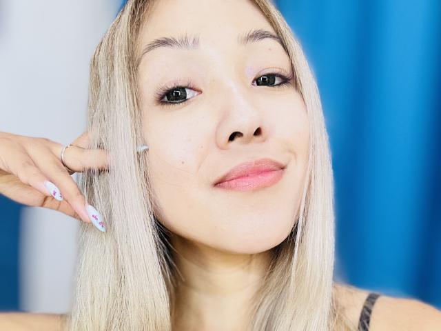 RenyLime with blonde hair, blue eyes, pink lipstick, and light skin, smiling slightly, having a hand with long, white nails touch her cheek, blue background.