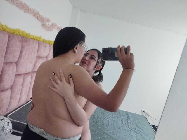 KatAndCharlotte with black hair and glasses, topless, being hugged from behind by Charlotte with pigtails, taking a selfie. Both have visible tits, wearing only gray underwear.