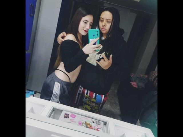 KatAndCharlotte with long brown hair, off-shoulder black top, exposing small tits, and gray shorts, stands next to Charlotte with black hair, hoodie, and colorful pants, taking a mirror selfie.