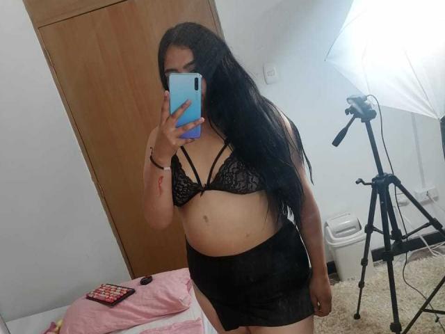 SexyHally with long black hair, wearing a black lace bra and black sheer skirt, holding a blue phone, standing in a room with a tripod and white light.