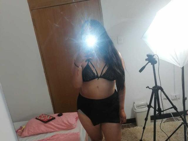SexyHally with long dark hair, black lace bra, and black high-waisted shorts, taking a mirror selfie with a flash-covered camera in a room with a tripod and bed.
