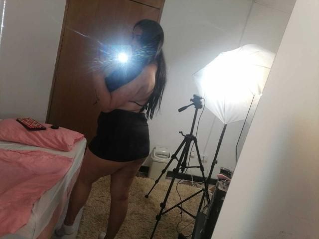 SexyHally with long dark hair, wearing a black top and shorts, showing her bare ass in a mirror selfie, flash from camera, standing in a room with a tripod.