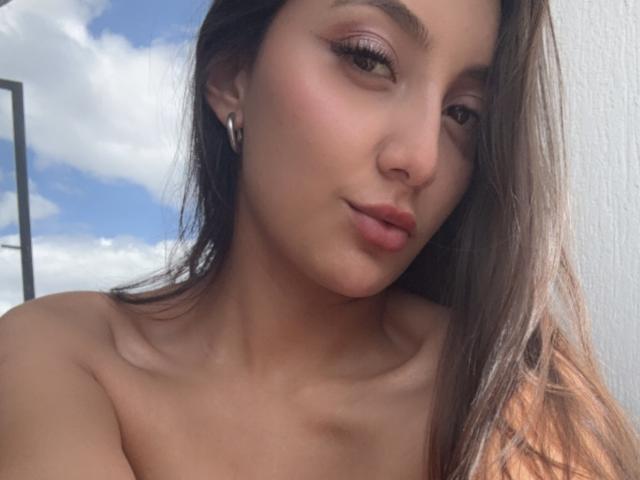 DianaFernandezz with pouty lips, brown eyes, and long brown hair, wearing no clothes, showing her bare shoulders and earrings, under a blue sky with white clouds.