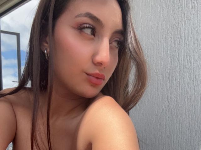 DianaFernandezz has brown eyes, subtle makeup, and pink lips, with long brown hair and a slight smile, showing bare shoulders, wearing a silver earring, in a relaxed selfie.