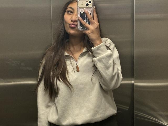 DianaFernandezz with long brown hair, tongue out, wearing a beige zip-up hoodie, holding a phone with a floral case, in a metal elevator. Boobs not visible.