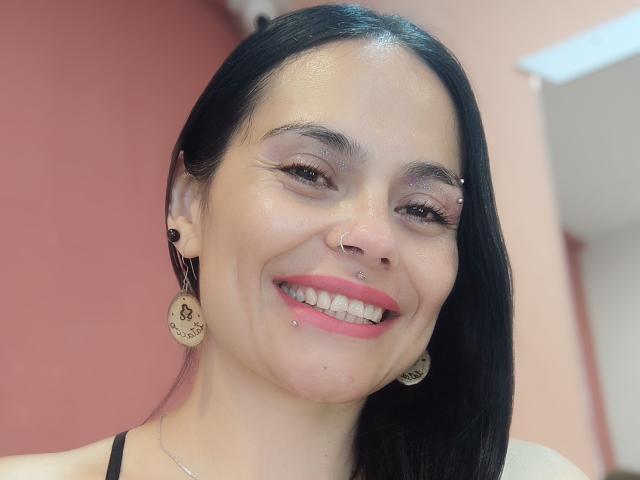 CharlotteCrofort has black hair, pink lipstick, nose ring, and lip piercing, smiling with visible teeth, wearing circular earrings, and has a slight tan with pinkish skin.