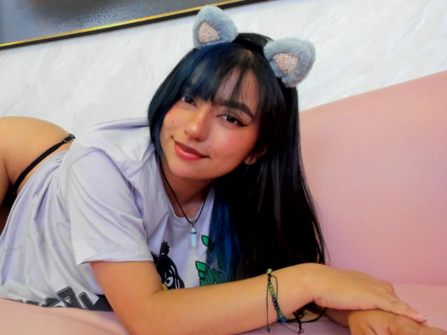 SussiieConor, with cat ears, laying on pink couch, smiling, wearing white shirt, black thong, blue-tipped black hair, bracelet, and nose ring, showing her ass, cute, cute face.