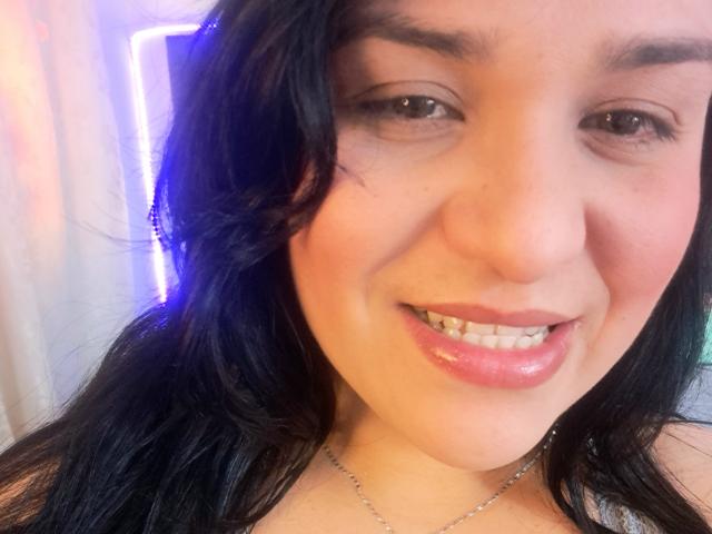 VivianaThompson's face, smiling with glossy pink lips, dark wavy hair, brown eyes, and a silver necklace, showing her teeth and slightly blushing. Her ass is not visible.