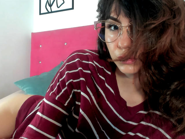 KariCarter with curly brown hair, wearing glasses and a maroon striped shirt, lying on a bed, showing a pouty face, with her right ass cheek partially visible.