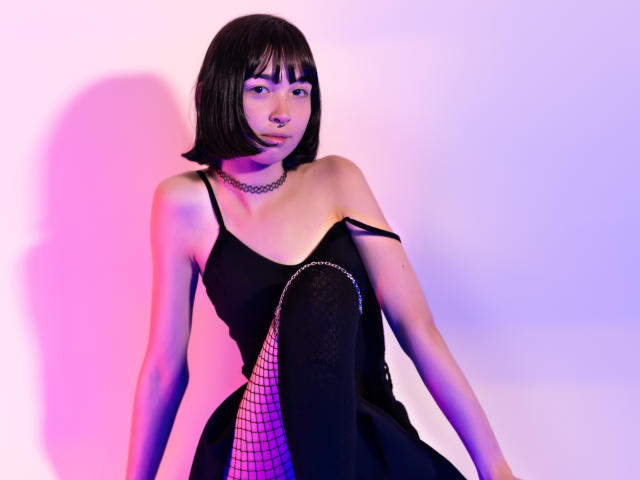 MillaHottLove69 with short black bob, black fishnet dress, black choker, and pink-purple lighting. She's pouting, with one strap off her dress, showing her small tits.