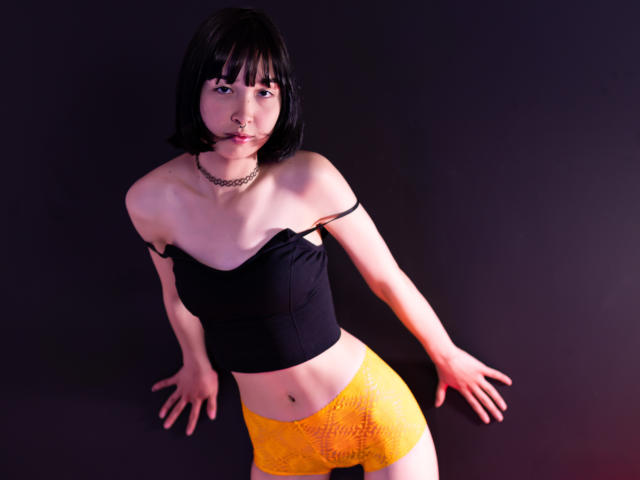 MillaHottLove69 with black crop top, yellow lace panties, black bob, chain necklace, and ball gag, leaning against dark background, arms spread, looking at camera.