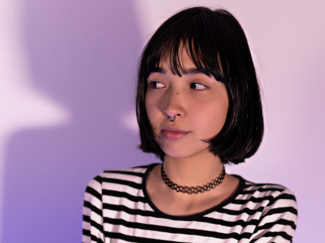 MillaHottLove69 ha un caschetto nero, un anello al naso, una collana e una camicia a righe, e guarda di lato con un leggero sorriso. È carina, con un naso piccolo e una pelle chiara..