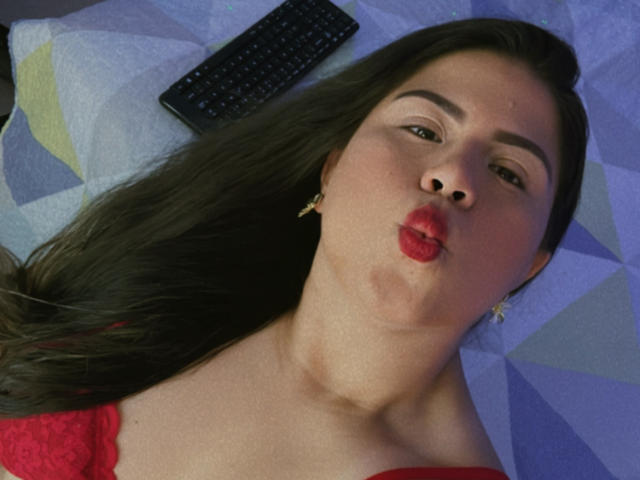 LaliForest with puffy red lips, dark brown hair, and gold earrings, lying on a bed with a black remote control, wearing a red lace top, making a kissy face.