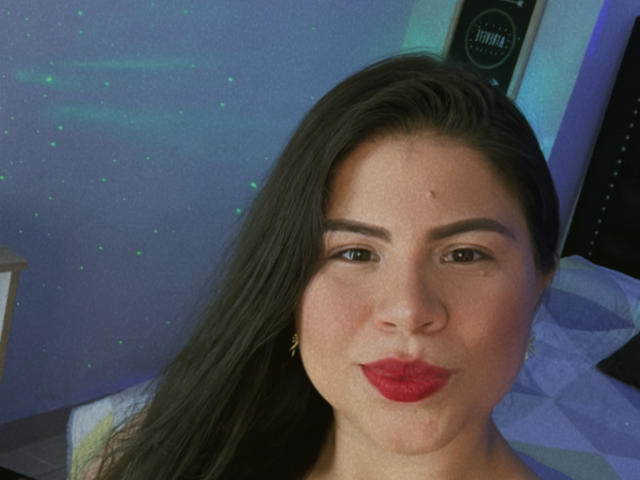 LaliForest has dark brown hair, red lipstick, and is wearing small gold earrings. She's looking at the camera with a subtle smile. Her skin is light, and she's slightly chubby.