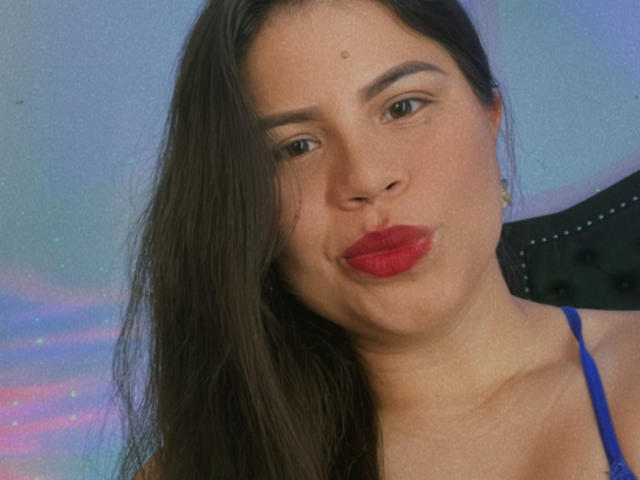 LaliForest has long brown hair, red lipstick, and a small mole above her lip. She's pouting her ass, wearing a blue spaghetti strap top, against a soft blue background.