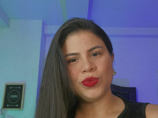 LaliForest, con capelli scuri, rossetto rosso e un leggero sorriso, indossa una camicia nera, guarda la fotocamera con un sorriso appena accennato, sfondo di una stanza blu, leggermente sfocato, luce soffusa..