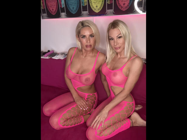 Two FoxyLynn-hot women with blonde hair, pink fishnet bodysuits, and visible tits, sitting on a pink couch, legs crossed, hands on knees, looking at camera.