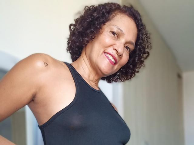MatureJennyX with curly black hair, smiling, wearing a black tank top, showing a small heart tattoo on her right shoulder, her boobs slightly visible through the fabric.