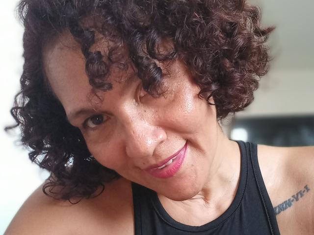 MatureJennyX has curly dark brown hair, a slight smile, and pink lipstick. She's wearing a black tank top, showing a tattoo on her right shoulder. Her skin is glistening.