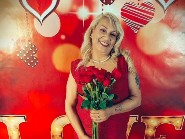 MatureYayita with blonde hair, red dress, pearl necklace, holding red roses, smiling, tattoo on right arm, red background with hearts and bokeh effect, standing, looking cute.