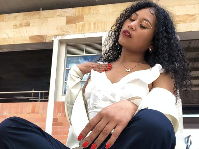 SophieSonyas with curly black hair, gold eyeliner, red lipstick, white top, black pants, red nails, white jacket, and butterfly necklace, sitting, hand on chest, looking down.