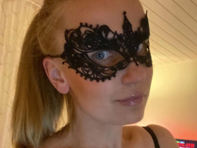 Leilani69, met blauwe ogen, draagt een zwart kanten masker, heeft glanzende, roze lippen, een blond staartje en laat haar borsten zien op de foto. Haar gezicht is lichtjes gedraaid..