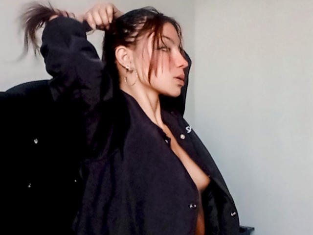 Ashley Miller, com cabelo castanho escuro em um coque bagunçado, vestindo uma camisa preta aberta, expondo seus pequenos seios, puxando o cabelo para cima com uma mão, em um perfil lateral..