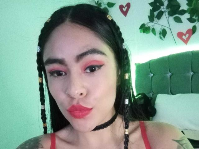 StellaBeckker with red eyeliner, pink lipstick, black braids, nose ring, and black choker, pouting lips, green headboard, and leafy wall decorations.