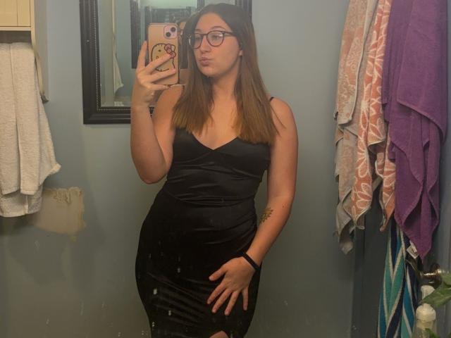 MaddieTheBaddie with long brown hair, glasses, black dress, and hand on hip, standing in bathroom, taking mirror selfie with phone, showing curvy ass and small tits.