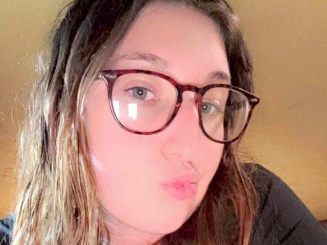 MaddieTheBaddie with glasses, pouty lips, nose ring, and messy brown hair, wearing a black top, looking into the camera.