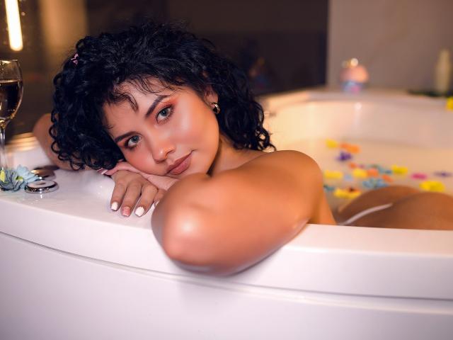 CanelaLebrad, con cabello negro rizado, piel morena y ojos verdes, se recuesta en una bañera blanca, con los brazos sobre el borde, rodeada de pétalos, mirando a la cámara con una ligera sonrisa..