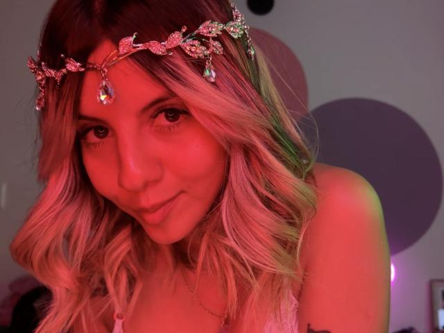 SweetAnaFlowers with pink and blonde wavy hair, wearing a sparkly flower crown, smiling at the camera, red light highlighting her face, visible cleavage in a low-cut top.