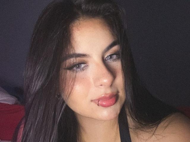 CharlotteBlass has long dark hair, blue eyes with thick eyeliner, and is sticking out her pink tongue with a small drop of white liquid on her chin.