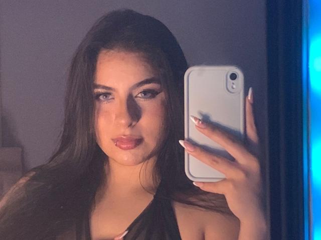 CharlotteBlass with long dark hair, full lips, and makeup, holding a white phone, taking a selfie, showing off her nails, wearing a black top, in dim lighting.