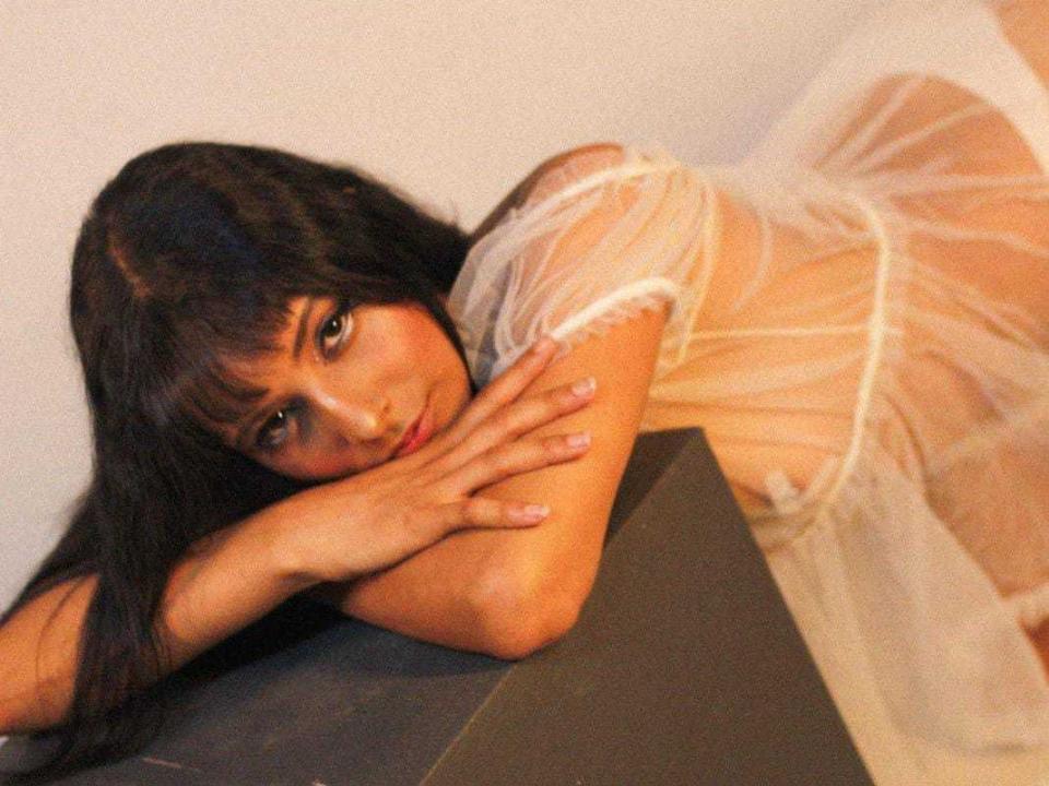 Liotta with long black hair, lying on a dark surface, wearing a sheer white dress, arms folded, hands near her face, looking at the camera with a subtle smile.