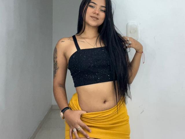 TamaraSummer with long black hair, wearing a black sparkly crop top, yellow skirt, and rings, standing in a hallway, touching her skirt with a confident smile.