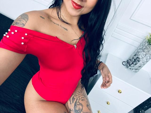 TamaraSummer with long black hair, red off-shoulder top, showing her big ass and tattoos, wearing red lipstick, holding phone, in a white room with black floor.