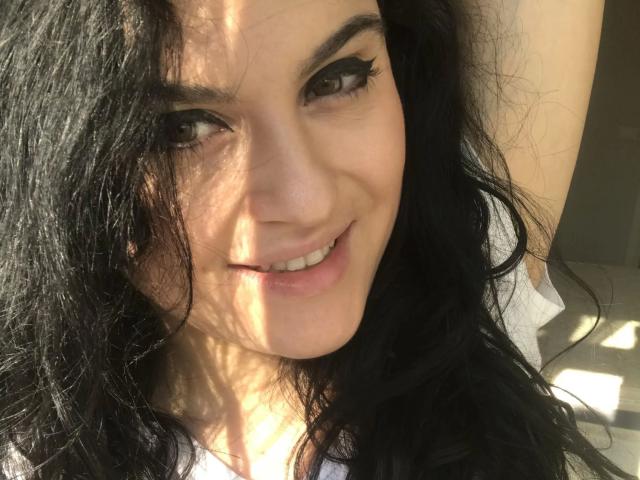 GeileMelly-hot with black hair, smiling, sunlit face, long eyelashes, white shirt, arm raised, light shadow on cheek, natural light, soft smile, cute, attractive, light skin, relaxed.