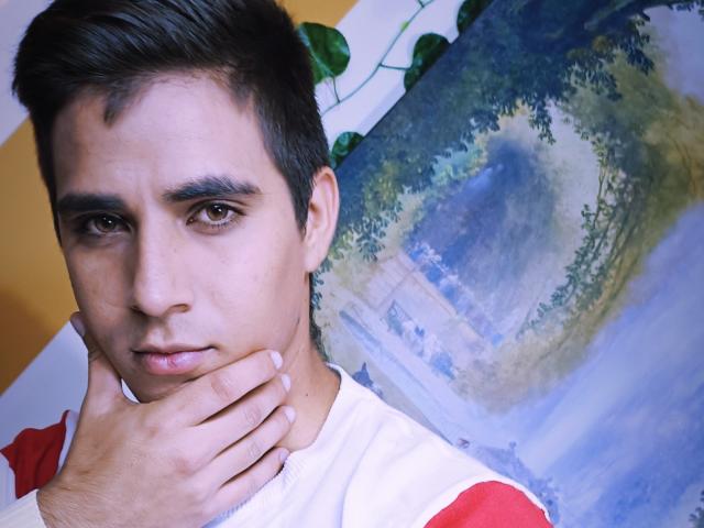 SantosMontenegro with dark hair, brown eyes, and a serious face, holding his chin with his left hand. Wearing a white and red shirt. Background has a painted landscape.