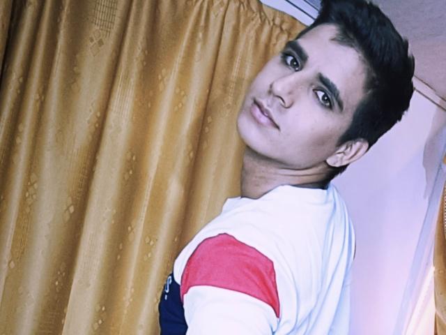 SantosMontenegro with short black hair, brown eyes, and a white shirt with red and blue sleeves, looking over shoulder, standing in front of golden curtains.