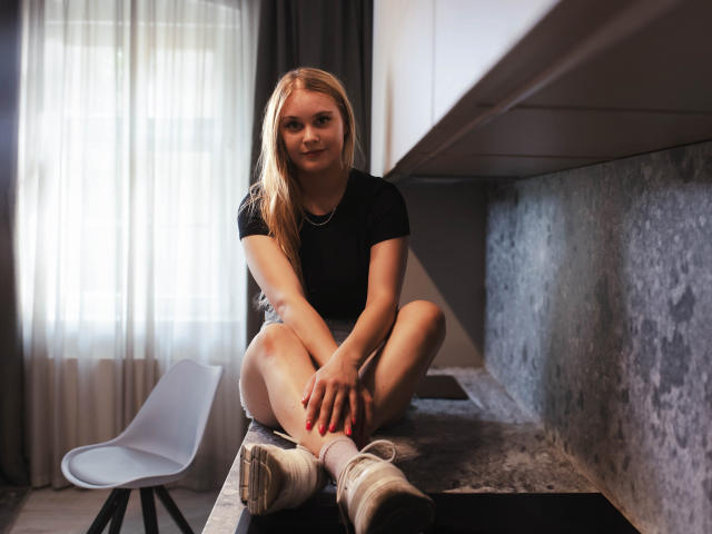ValeriaDream, blonde, sitting on kitchen counter, legs crossed, black shirt, white socks, sneakers, red nails, subtle smile, arms resting on knees, gray textured wall, white chair.