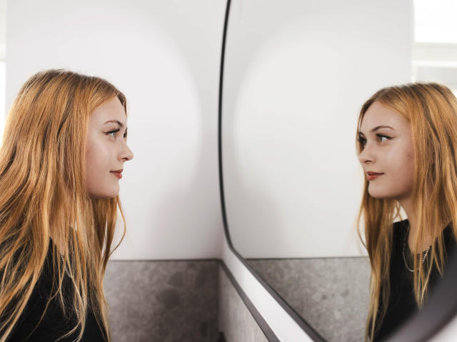 ValeriaDream with long blonde hair, looking at her reflection in a mirror. Her face is serious, she's wearing a black top, and her reflection is perfectly mirrored.