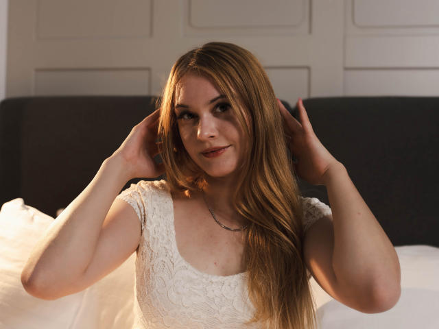ValeriaDream with long, blonde hair, wearing a white lace dress, hands in her hair, smiling softly, sitting on a bed with white pillows, warm light on her face.