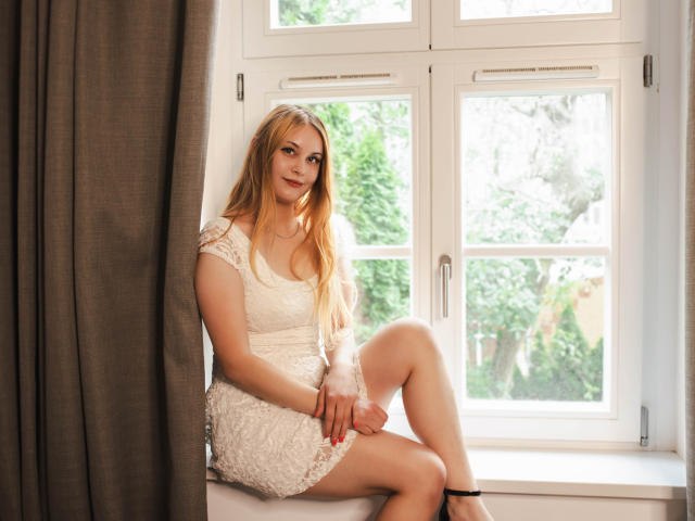 ValeriaDream with long blonde hair, white lace dress, and black heels, sitting on a window sill, legs crossed, looking at camera, soft smile, daytime, brown curtain to left.