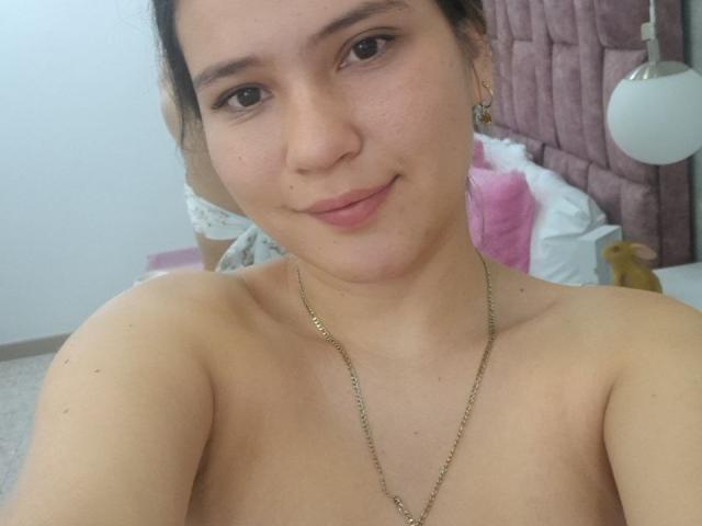 CelesteDash with light skin, brown eyes, and small tits, smiling at the camera, wearing a gold necklace, and no top, in a bedroom with pink and white bedding.