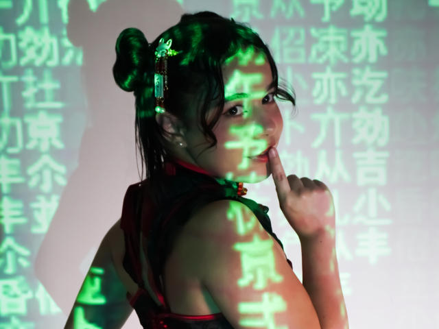 LissyCarter with dark hair in buns, wearing a red and black Chinese dress, smiling, finger touching her lip, green Chinese characters projected on her.