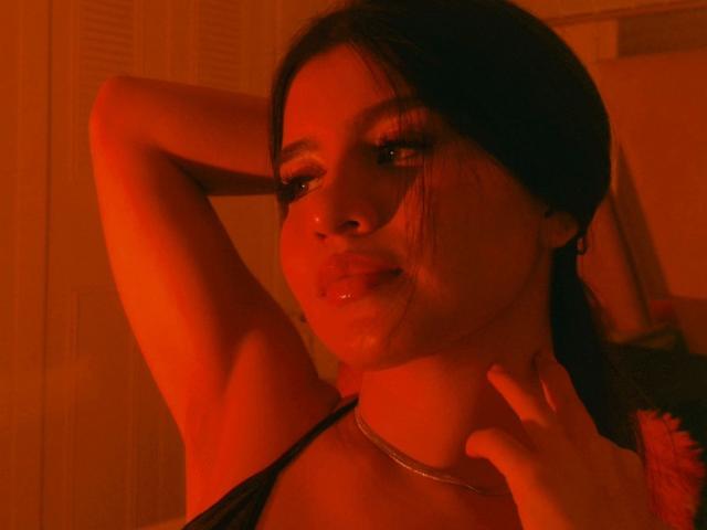 SaraLens with red-lit skin, dark hair in pigtails, hand behind head, necklace, glossy lips, slightly open mouth, visible wetness on cheek, black strap, sexy, seductive expression.