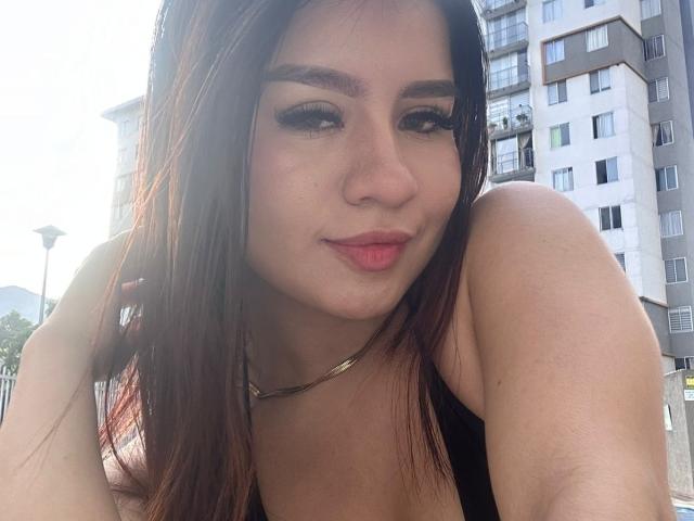 SaraLens with brown hair, black eyeliner, and pink lipstick, wearing a black top, her ass slightly visible, sunlight on her face, hand in hair, subtle smile.