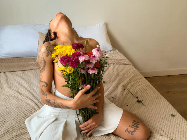 AngelitaFlor, senza top, con tatuaggi, che tiene un bouquet di fiori gialli, rosa e viola, è sdraiata su un letto, indossa una gonna bianca, con la testa inclinata verso l'alto, su un pavimento di legno, con un cuscino bianco..